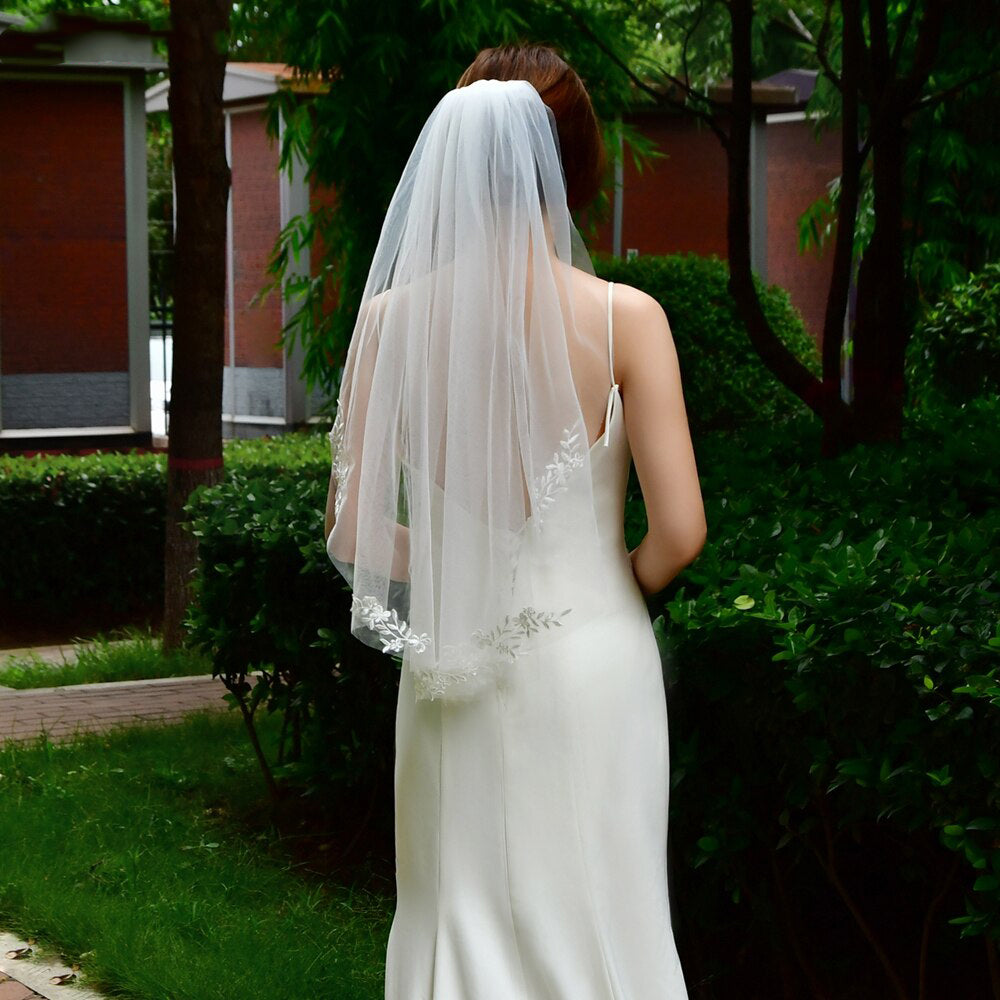 A bride in a white dress and the Bergamot Bridal fingertip length embroidered lace applique veil standing in a garden, facing away from the camera, surrounded by green foliage, possibly contemplating her choice from one of the bridal shops in London Ontario.