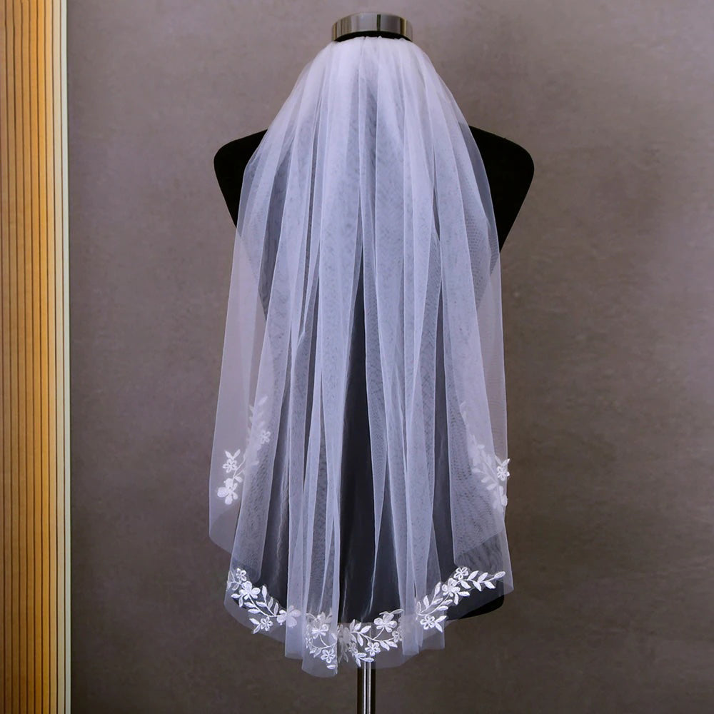 A Fingertip length embroidered lace applique veil by Bergamot Bridal displayed in a bridal shop against a wooden panel background.