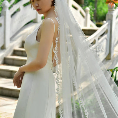 A bride in a white sleeveless gown from one of the bridal shops in London, with a long, Bergamot Bridal delicate embroidered leaf lace trimmed veil stands alongside a stone staircase in a lush garden.