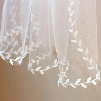 White sheer curtains with a Bergamot Bridal delicate embroidered leaf lace trimmed veil, perfect for bridal shops, hanging against a light background.
