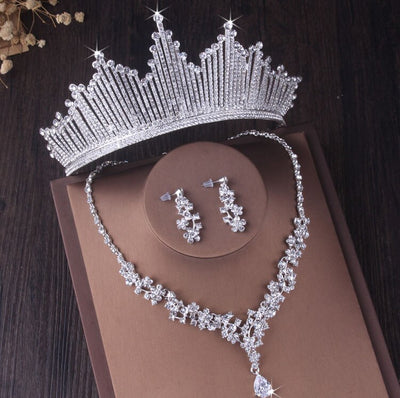A sparkling Bergamot Bridal Silver Crystal Bridal Jewelry Set, including a necklace, earrings, and tiara, displayed on a brown book against a dark wooden background.