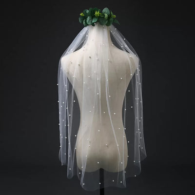A Bergamot Bridal ivory pearl beaded bridal veil displayed on a mannequin head in a London bridal shop, adorned with a green floral crown against a dark background.