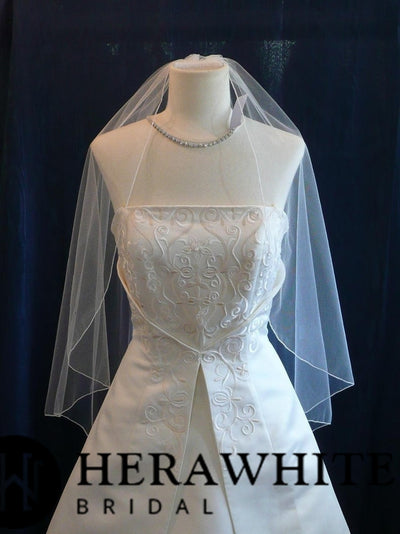 A bridal gown on a mannequin, featuring an embroidered bodice with floral patterns, a pleated skirt, and an Ivory Single Tiered Bridal Veil by Bergamot Bridal. The label "HERAWITE BRIDAL" is visible at the bottom of the image.