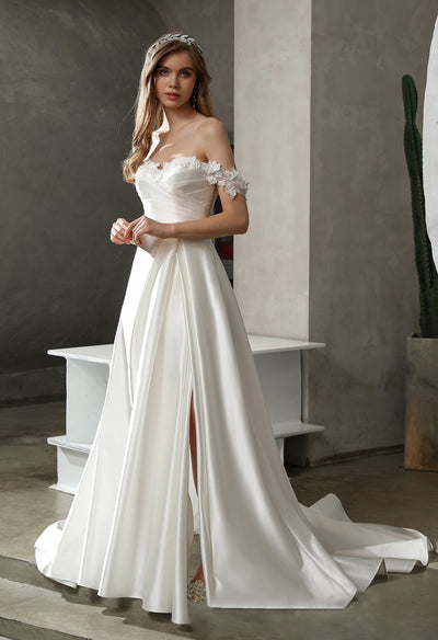 A bride in an elegant off-the-shoulder wedding gown with a train, standing in a rustic indoor setting.
