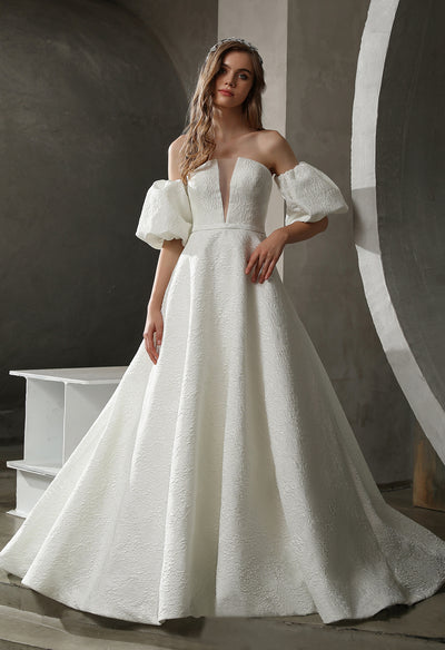 A woman in an elegant white off-shoulder gown with a plunging neckline, posing in a room with minimalist gray arches.