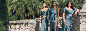 Three women in matching teal dresses standing in a lush garden with palm trees.