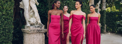 Four women in matching pink dresses standing beside a classical statue in a lush garden setting.