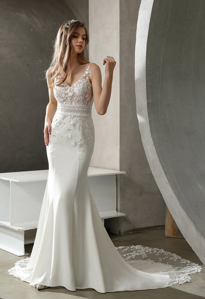 A woman in an elegant white wedding dress with lace details stands in a minimalist room, looking thoughtfully to the side.