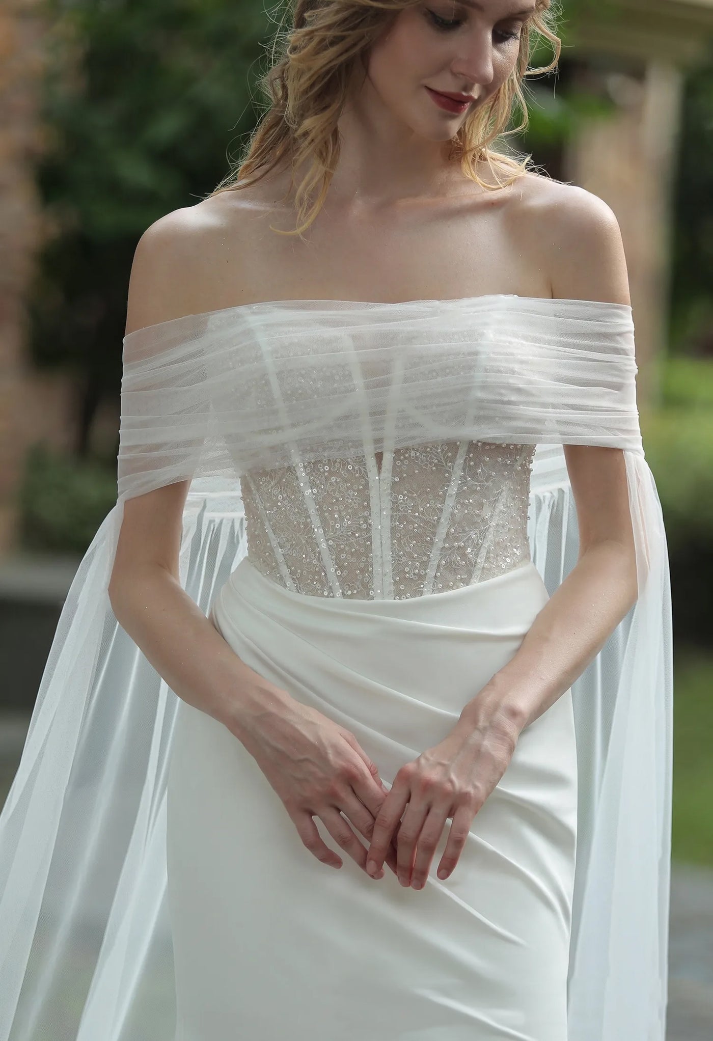A woman in an elegant off-shoulder Bergamot Bridal wedding dress with a beaded bodice and sheer overlay, gracefully adjusting the fabric near her waist.