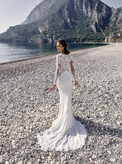 A bride in a EddyK Isandra gown from Bergamot Bridal with a sweetheart neckline stands on a pebble beach with towering cliffs and calm sea in the background.