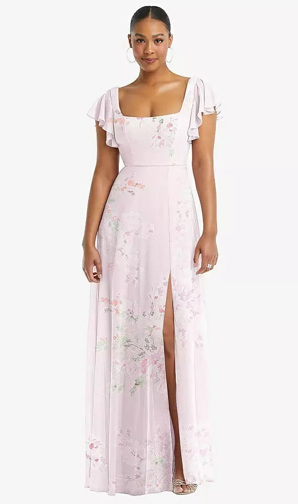 A woman wearing the Flutter Sleeve Scoop Open-Back Chiffon Maxi Bridesmaid Dress - Style 1568 by Dessy from Bergamot Bridal stands against a white background, its light pink floral pattern, flutter sleeves, thigh-high slit, and square neckline adding an elegant touch.