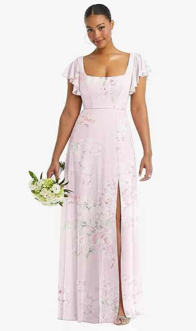 A woman in the Flutter Sleeve Scoop Open-Back Chiffon Maxi Bridesmaid Dress - Style 1568 by Dessy from Bergamot Bridal, featuring a light pink floral pattern, flutter sleeves, and a thigh-high slit, holds a small bouquet of white flowers.