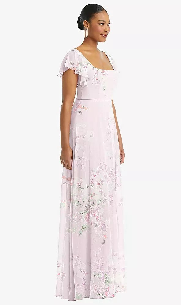 A woman in a Flutter Sleeve Scoop Open-Back Chiffon Maxi Bridesmaid Dress by Bergamot Bridal, style 1568, light pink in color, with hoop earrings is standing on a plain background.