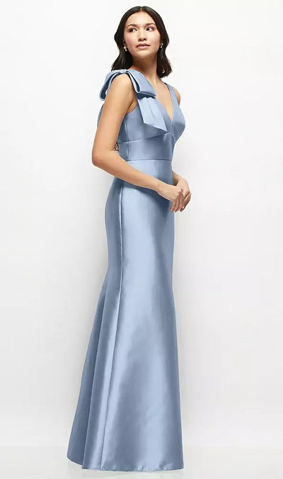 A woman elegantly models the Bergamot Bridal's Deep V-back Satin Trumpet Bridesmaid Dress - Style D863 by Dessy, showcasing its floor-length, sleeveless design with a stunning handcrafted cascading bow at one shoulder, set against a simple backdrop.