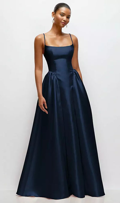 A woman in a stylish navy satin maxi dress, featuring a scoop neck and drop basque skirt, stands gracefully against a plain white background. The dress, from Bergamot Bridal's Dessy Collection Jillian (Style 3169), includes thin straps and pockets.