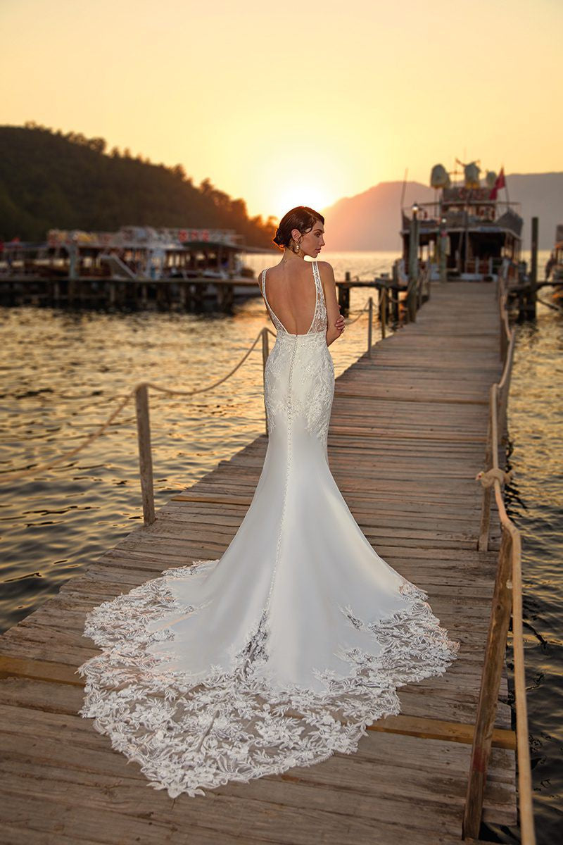 A bride in an Eddy K Arianna Mermaid Wedding Gown from Bergamot Bridal, with a beaded lace bodice, standing on a wooden pier at sunset, looking towards a mountainous horizon.