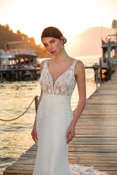 A woman in an elegant Eddy K Arianna mermaid wedding gown from Bergamot Bridal, with a beaded lace bodice, standing on a dock at sunset, with tranquil water in the background and soft sunlight.