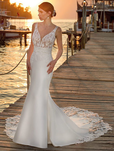 A woman in an elegant white Eddy K Arianna Mermaid Wedding Gown with beaded lace bodice stands on a wooden pier at sunset.