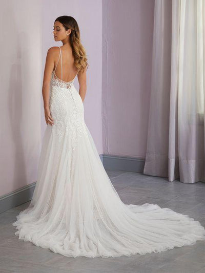 A woman in a Beaded Lace Fit & Flare Gown with Double Spaghetti Straps by Christina Wu, available from Bergamot Bridal, stands in a room with light-colored walls and curtains.