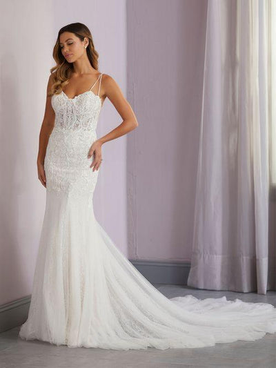 A woman elegantly stands near a wall and curtain in a Beaded Lace Fit & Flare Gown featuring double spaghetti straps and a tulle train by Christina Wu, offered by Bergamot Bridal.