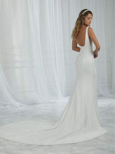 A woman in a Square Neckline Fitted Wedding Dress by Christina Wu, crafted from textured crepe and featuring a cathedral train, stands on a marble floor in front of sheer curtains.