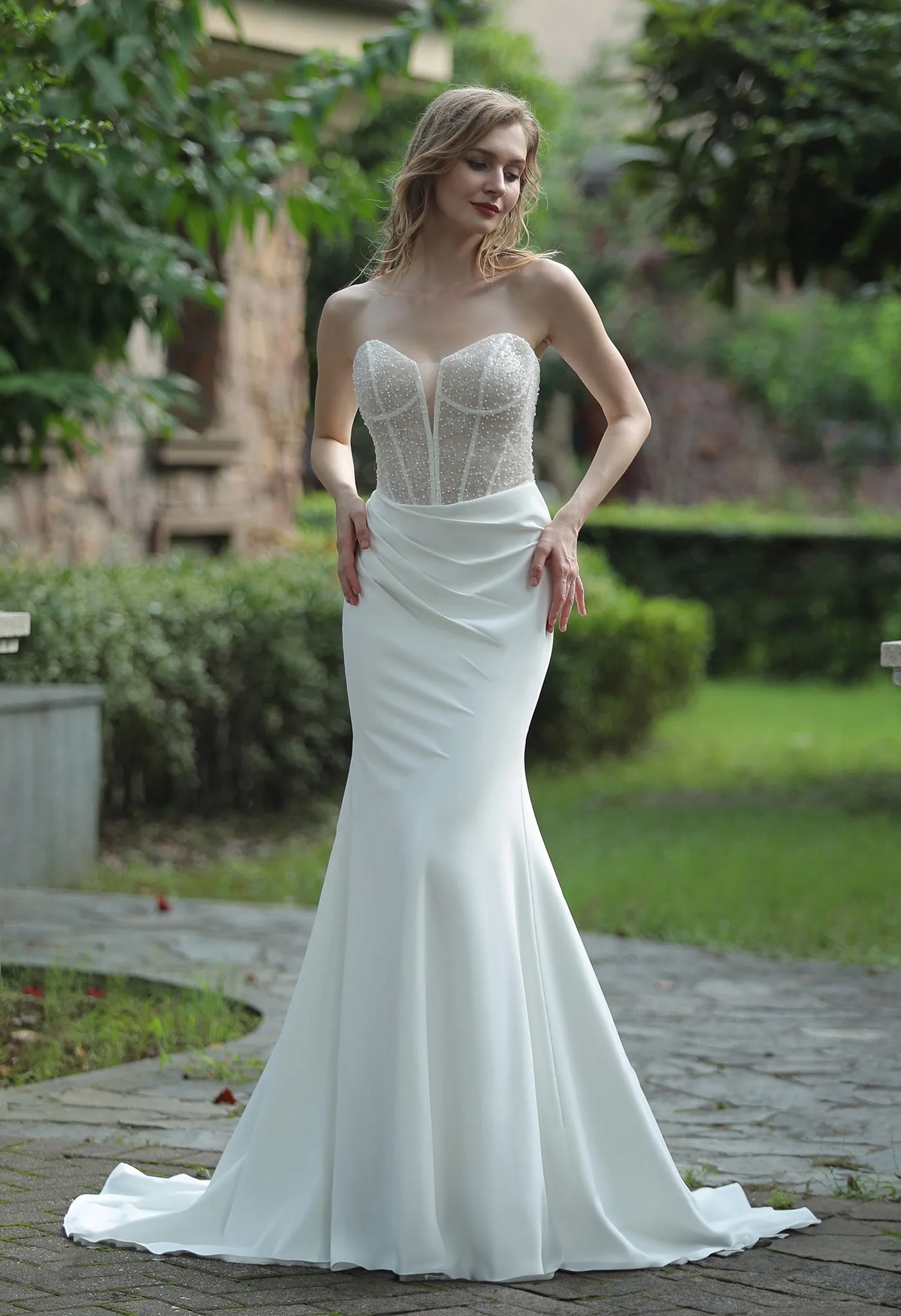 A woman in a Bergamot Bridal plunging sweetheart neckline beaded crepe fit and flare wedding dress with a fitted bodice and mermaid silhouette, posing outdoors.