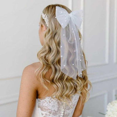 A woman with long, wavy blonde hair is seen from behind, wearing an Off-White lacy dress and the Bergamot Bridal "Bridal Hair Bow with Pearl Accents," a sheer bow resembling a delicate veil.