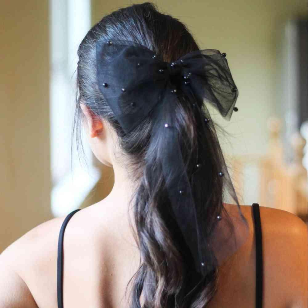 A woman with long, dark hair in a ponytail is shown from the back, wearing Bergamot Bridal’s “Bridal Hair Bow with Pearl Accents.”.