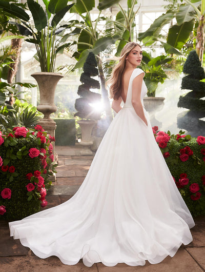 A woman in a white Mikaella 2253 Simple Bridal Ball Gown - Off The Rack stands in a sunlit greenhouse surrounded by lush plants and vibrant red flowers from Bergamot Bridal.