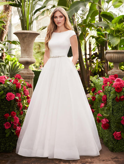 A woman stands in a lush garden, wearing an elegant white wedding dress with a satin top, bejeweled belt, and voluminous organza skirt from Bergamot Bridal's Mikaella 2253 Simple Bridal Ball Gown - Off The Rack.