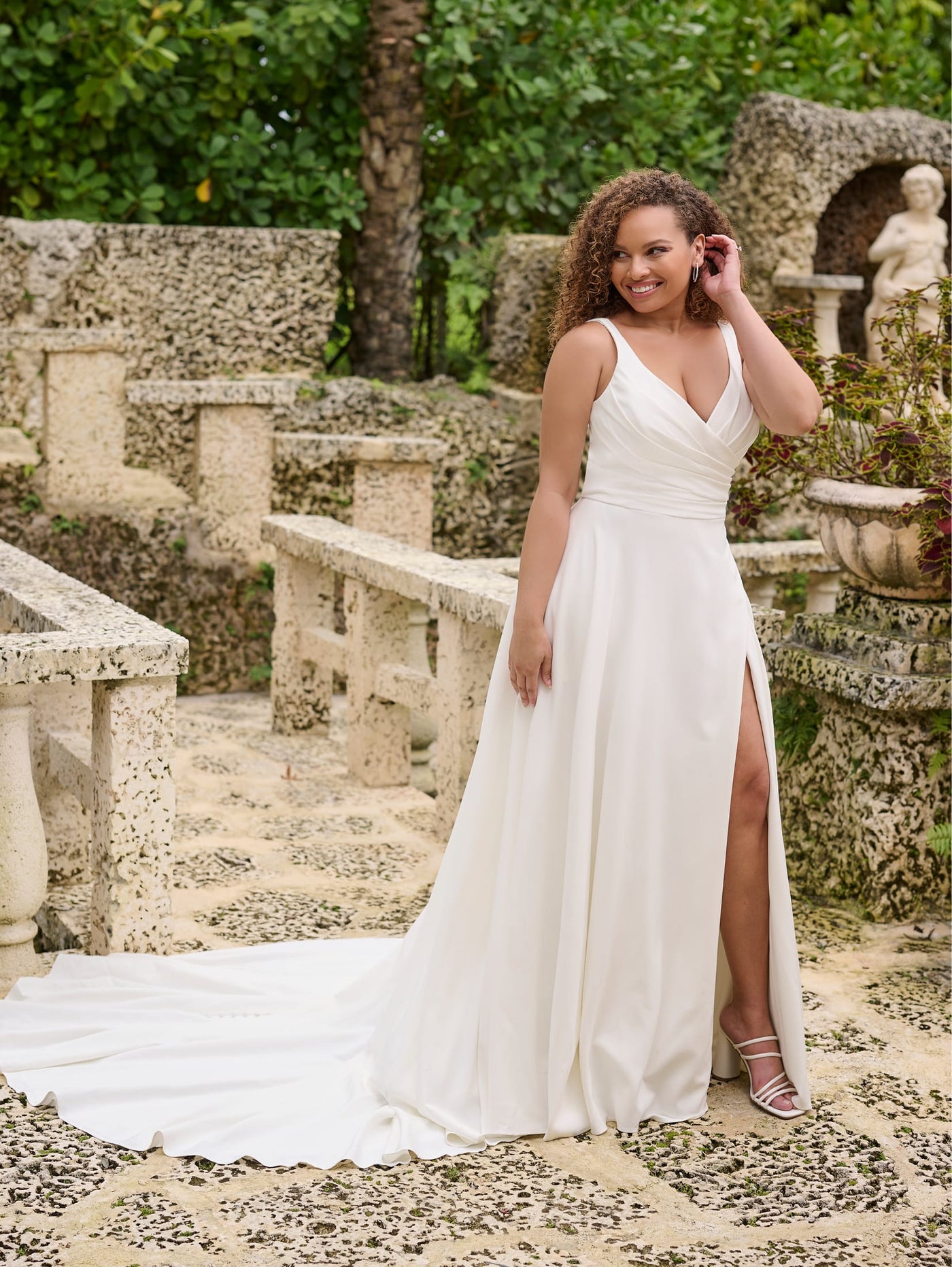 A woman in a Simple Curvy A-Line Wedding Gown with a ruched V-neckline bodice by Bergamot Bridal's Christina Wu stands on a stone pathway, surrounded by greenery and sculpted stonework.