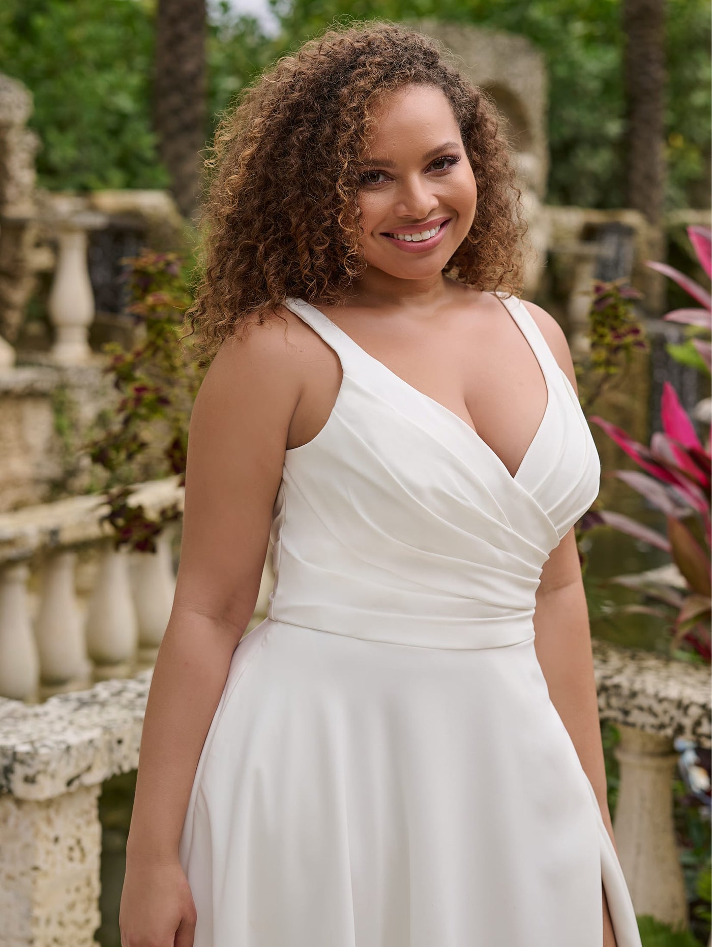 A woman wearing the Simple Curvy A-Line Wedding Gown with a Ruched V-Neckline Bodice by Christina Wu from Bergamot Bridal stands outdoors, smiling amidst lush greenery and stone railings.