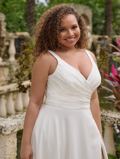 A woman wearing the Simple Curvy A-Line Wedding Gown with a Ruched V-Neckline Bodice by Christina Wu from Bergamot Bridal stands outdoors, smiling amidst lush greenery and stone railings.