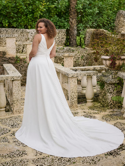 A woman wearing the Simple Curvy A-Line Wedding Gown with Ruched V-Neckline Bodice by Christina Wu from Bergamot Bridal gracefully poses outdoors in a garden, amidst stone structures and lush greenery.