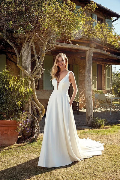A woman in a EddyK Madeline - EK 1355 - Off The Rack wedding dress with a plunging v-neckline smiling, standing outside a rustic house with greenery around.