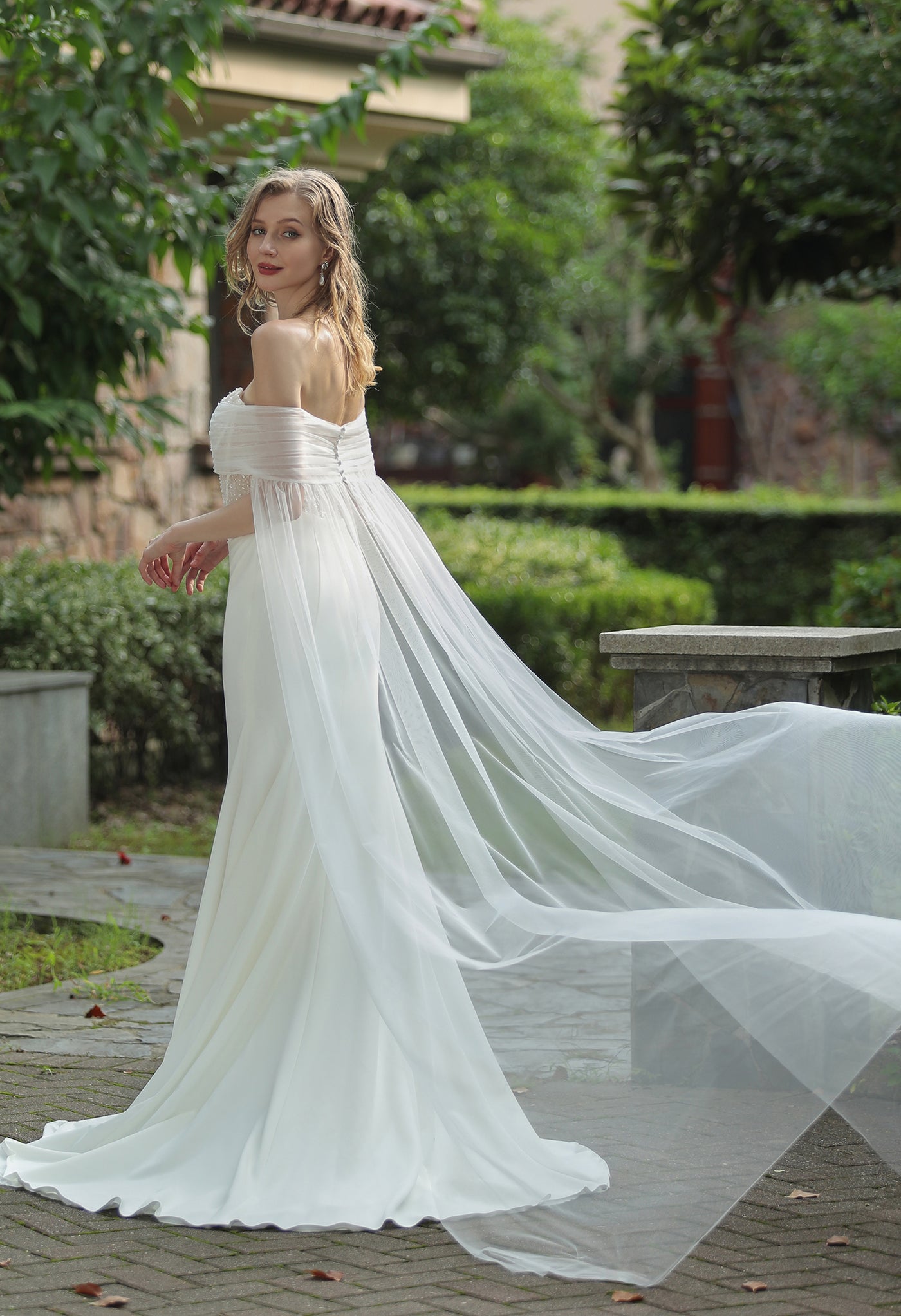A woman in a Bergamot Bridal plunging sweetheart neckline beaded crepe fit and flare wedding dress looks over her shoulder in a garden pathway, with scattered leaves and trees in the background.