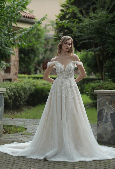 An Elegant Floral 3D Lace Wedding Dress With Off-Shoulder Straps from Bergamot Bridal standing in a garden.