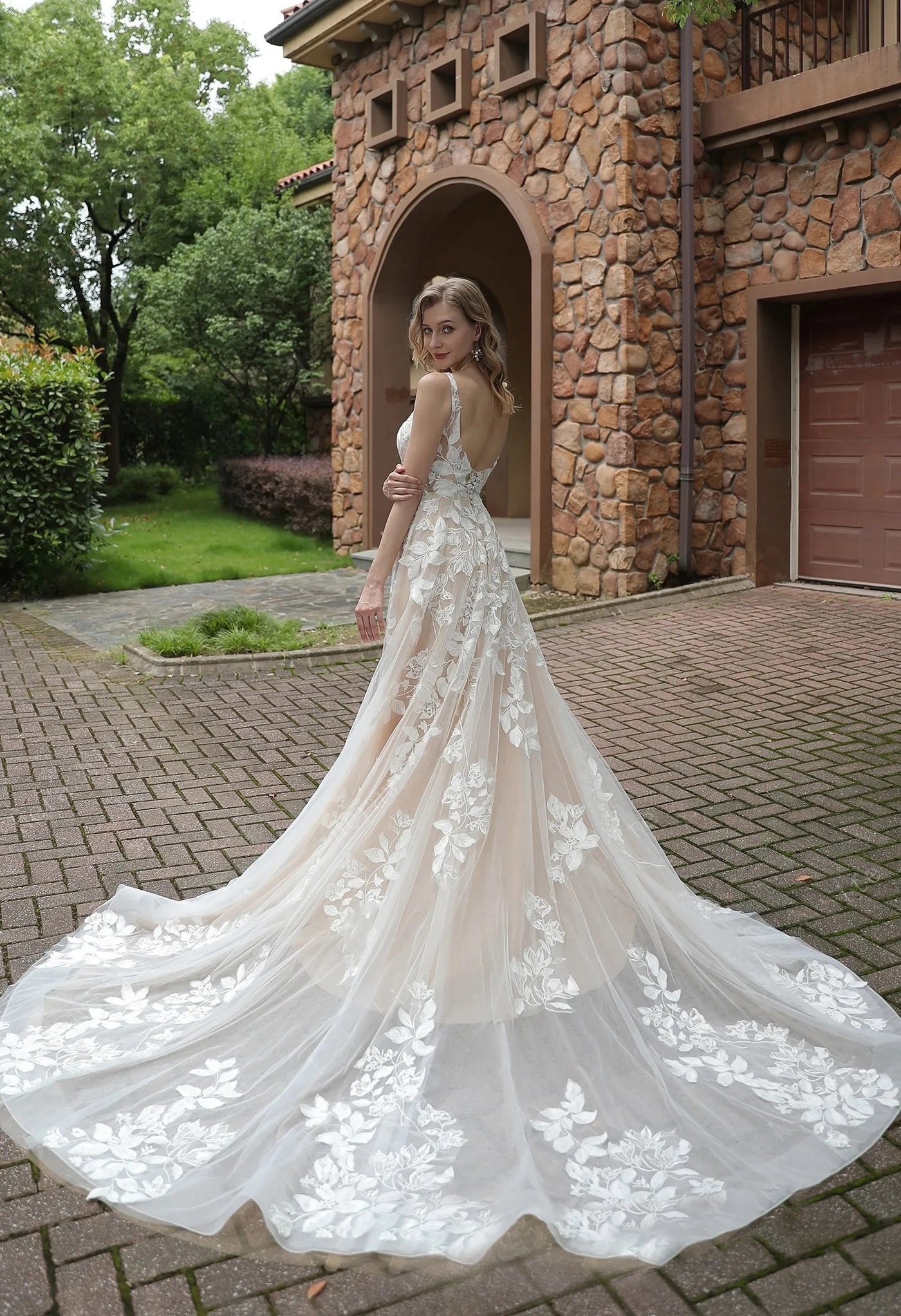A woman in a Bergamot Bridal Luxurious Floral Lace A-Line Wedding Dress With Sheer Train standing in front of a house.