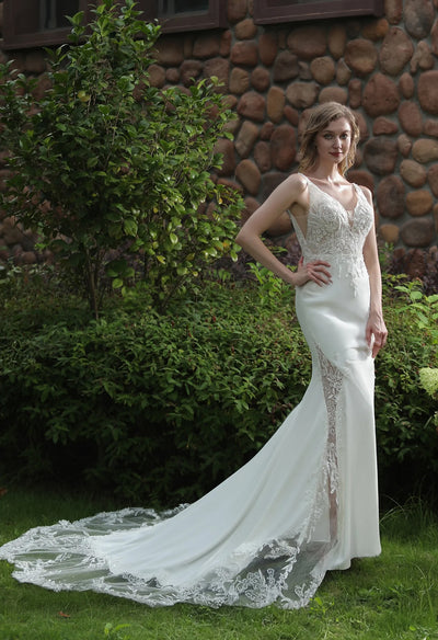 A person wearing a Bergamot Bridal Simple Beaded Fit And Flare Gown with V Neckline And Crepe Skirt, standing beside a green bush against a stone wall background.