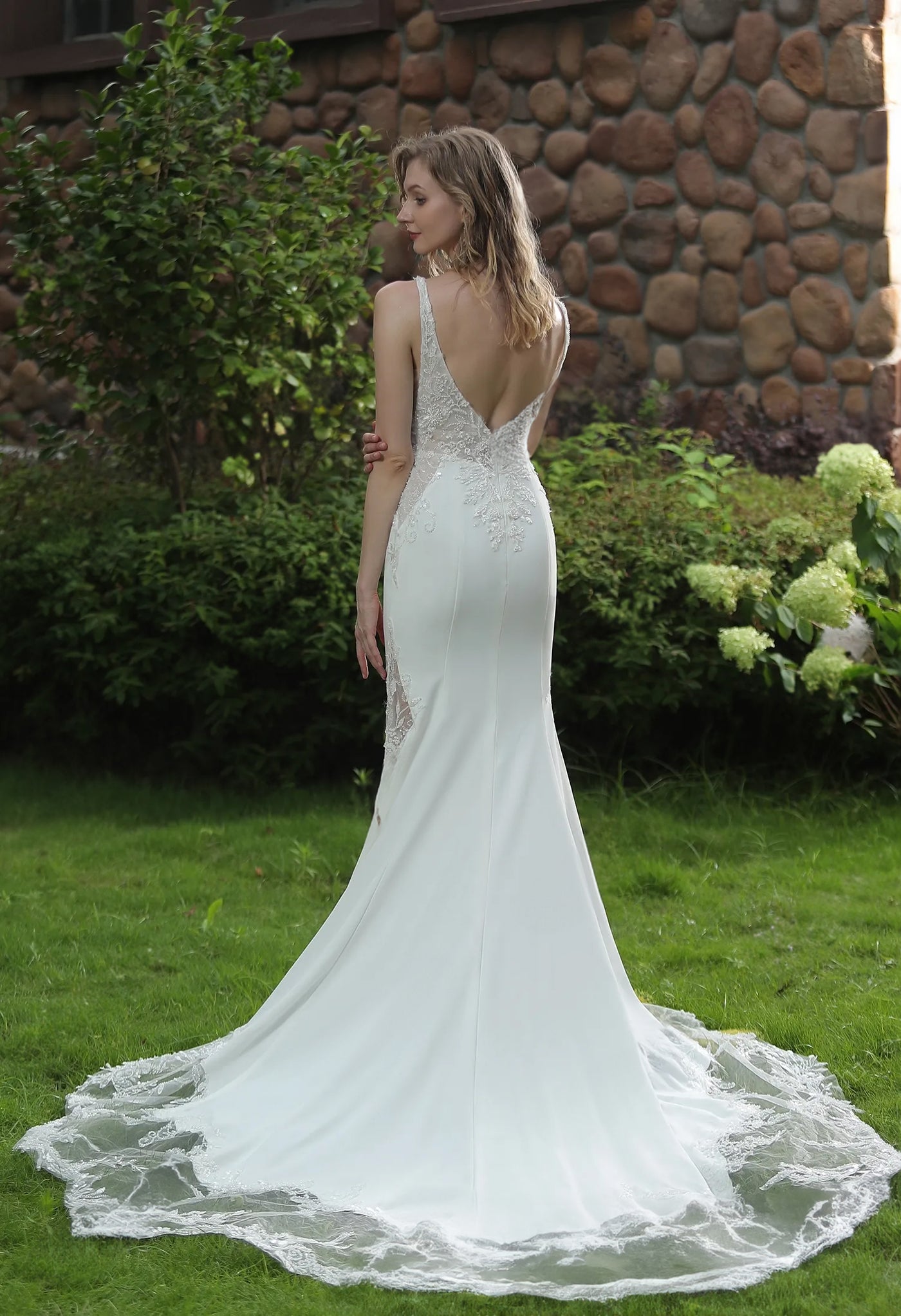 A woman standing on grass and facing away from the camera, wearing a Bergamot Bridal Simple Beaded Fit And Flare Gown with V Neckline And Crepe Skirt with an open back.
