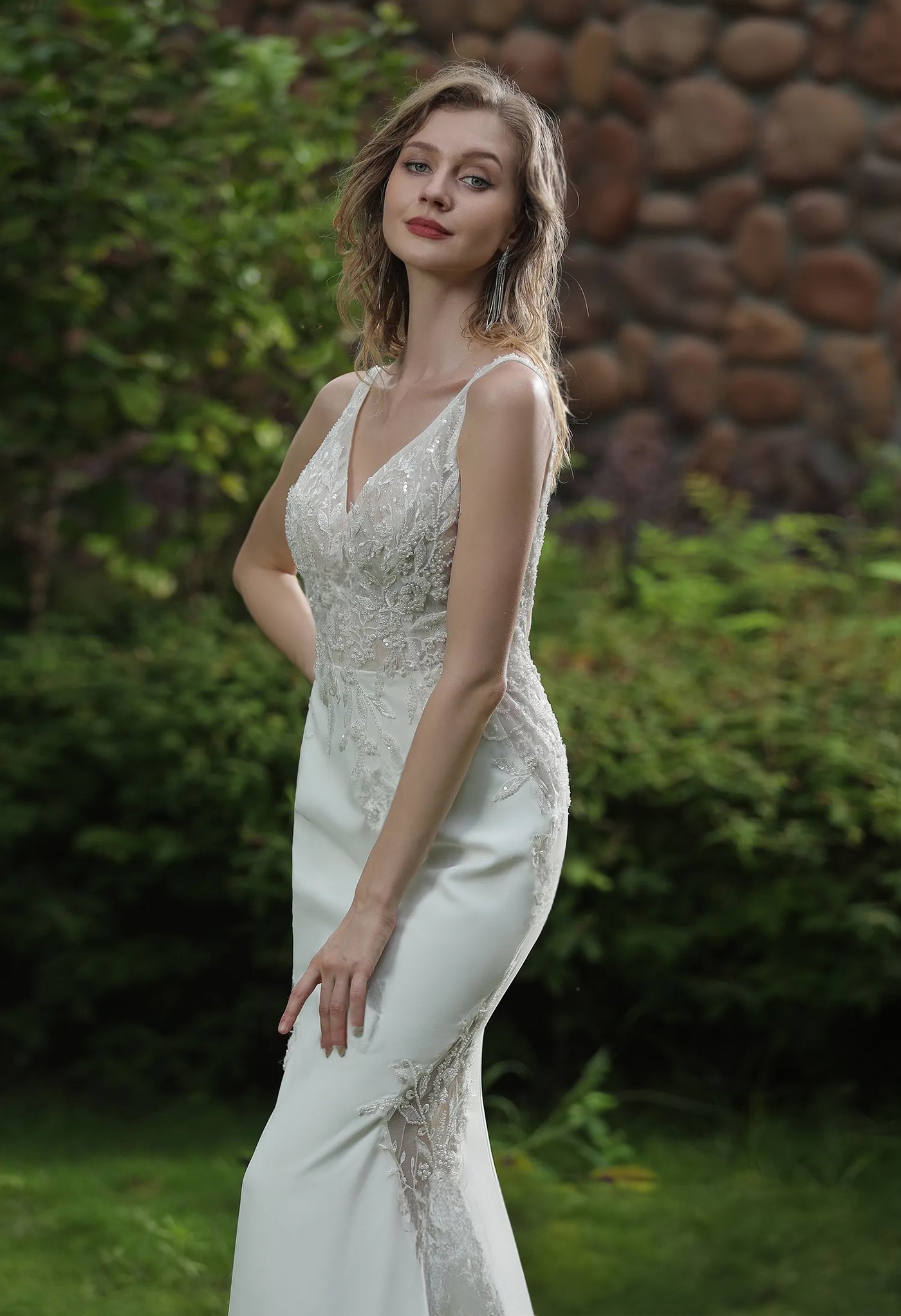 A woman in a Bergamot Bridal Simple Beaded Fit And Flare Gown with V Neckline And Crepe Skirt, adorned with beaded lace, posing outdoors with a natural background.