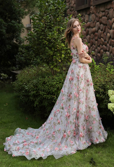 A woman in a pink and white floral wedding dress, the Romantic Square Neckline with 3D Flowers Bridal Gown With Detachable Sleeves by Bergamot Bridal, is posing in front of bushes.