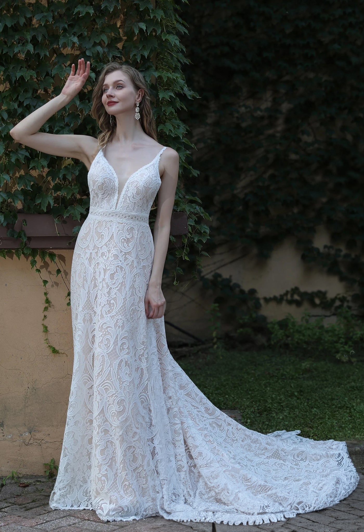 A bride wearing a Boho Lace Wedding Dress With Spaghetti Straps and Removable Bell Sleeves by Bergamot Bridal is posing in front of a brick wall.