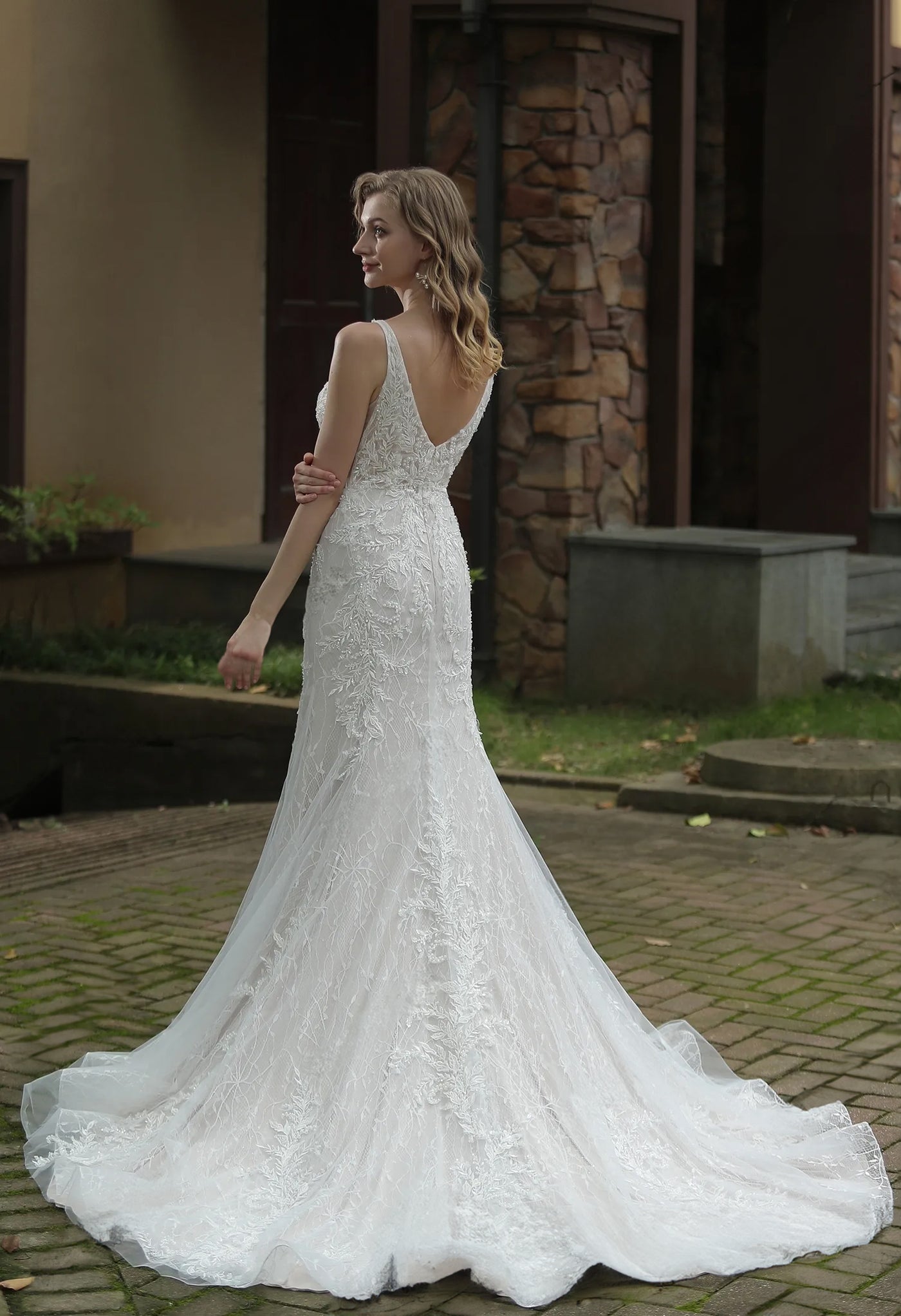 A bride in an elegant Bergamot Bridal Classic V-Neck Allover Lace Fit And Flare Wedding Dress with a train, posing outdoors.