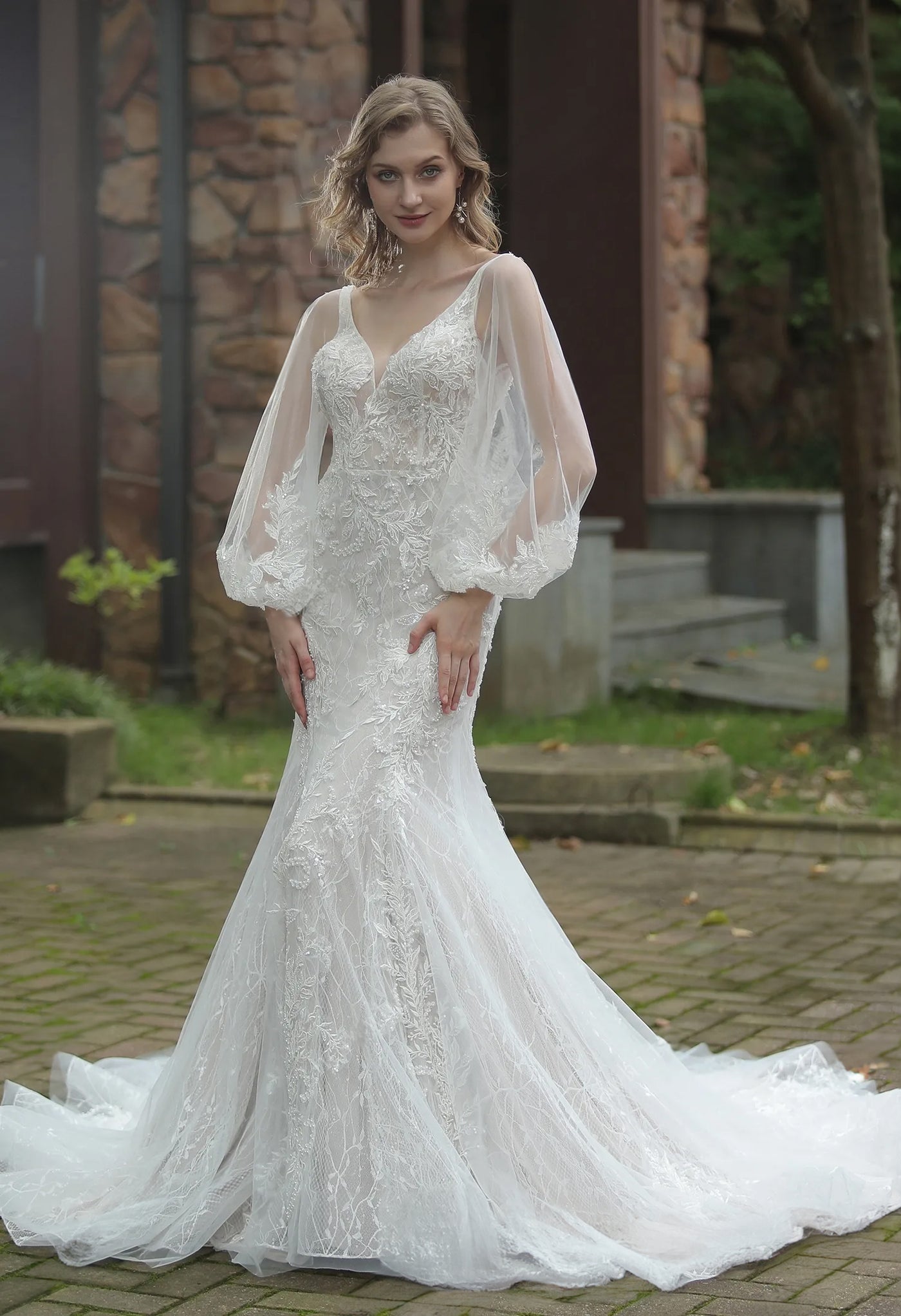 A bride in a Bergamot Bridal Classic V-Neck Allover Lace Fit And Flare Wedding Dress with a v-neckline and sheer sleeves poses outdoors.