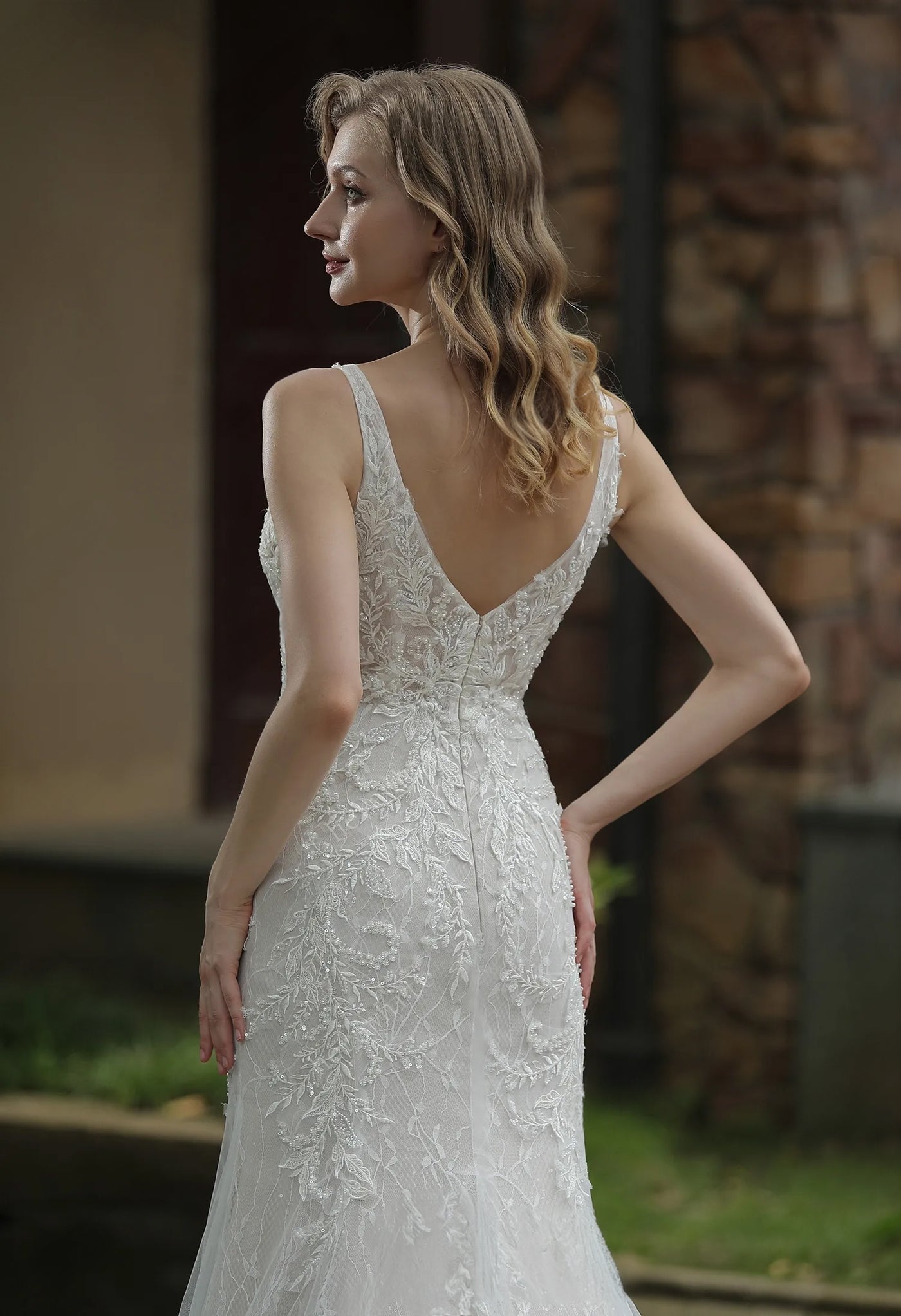 A woman wearing an elegant Bergamot Bridal Classic V-Neck Allover Lace Fit And Flare Wedding Dress gown looks over her shoulder.