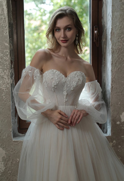 A beautiful bride in a Enchanting Pleated Tulle A-line Wedding Dress With Puff Sleeves by Bergamot Bridal posing in front of a window at a bridal shop.