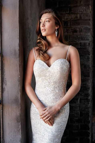 A woman in an elegant EddyK CT249 Beaded Sheath Wedding Gown - Off The Rack by Bergamot Bridal standing near a rustic wooden doorway, looking contemplatively to the side.