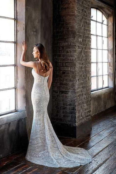 Woman in a EddyK CT249 Beaded Sheath Wedding Gown from Bergamot Bridal standing by a window in a rustic room, touching the window, looking outward.
