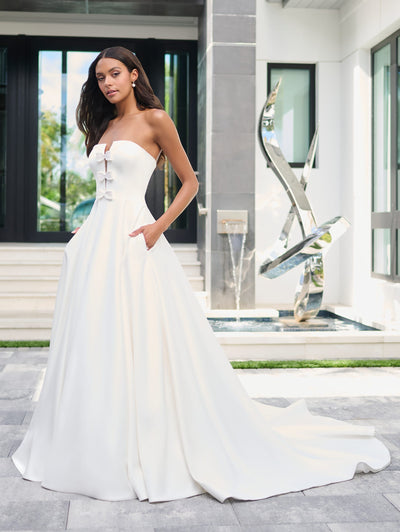 A woman wearing the Simple Satin Ball Gown with Pearl Bow Bodice Detail Wedding Dress by Adrianna Papell Platinum from Bergamot Bridal stands outdoors in front of a modern building with large windows and a metal sculpture.